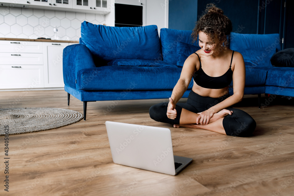 Happy beautiful woman show thumb up after finished online workout.Smiling young strong sporty fitness woman wearing black tracksuit doing yoga using laptop showing thumb up sitting on floor at home