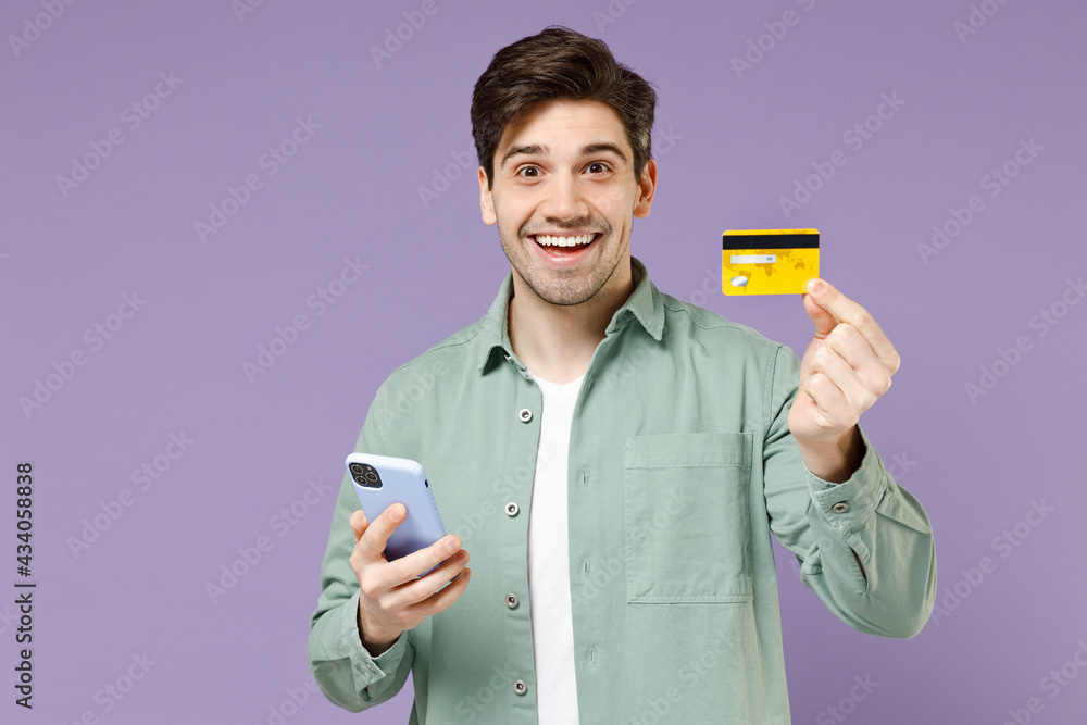 Young fun satisfied caucasian man 20s in casual mint shirt white t-shirt hold mobile cell phone credit bank card shopping online isolated on purple background studio portrait People lifestyle concept. Young fun satisfied caucasian man 20s in casual mint shirt white t-shirt hold mobile cell phone credit bank card shopping online isolated on purple background studio portrait People lifestyle concept.