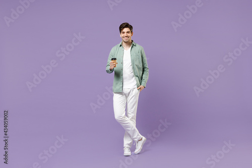 Full length young satisfied smiling fun man 20s wearing casual green mint shirt white t-shirt holding paper cup of coffee drink hot tea in morning isolated on purple color background studio portrait.