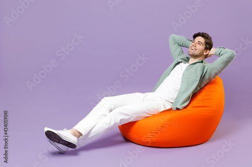 Full length fun man 20s in casual mint shirt white t-shirt sitting in orange bean bag chair hold hands folded behind neck isolated on purple color background studio portrait People lifestyle concept.