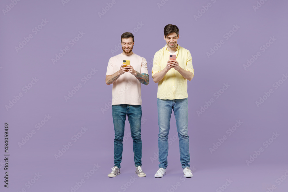 Full length fun smiling cool two young happy men friends together stand in casual t-shirt using mobile cell phone chat online isolated on purple background studio People friendship lifestyle concept Full length fun smiling cool two young happy men friends together stand in casual t-shirt using mobile cell phone chat online isolated on purple background studio People friendship lifestyle concept