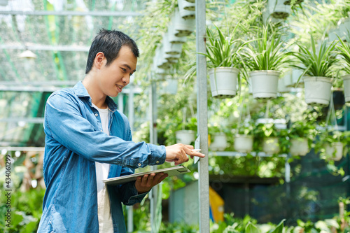 Smiling young man with clipboard in hands counting plants and flowers in gardening center