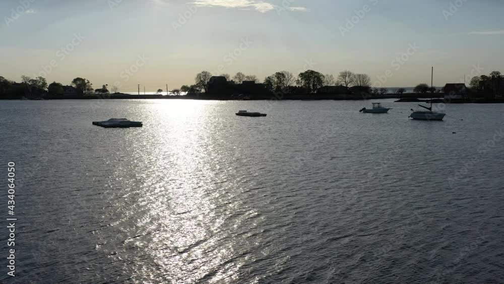 A low altitude drone shot over calm waters, as the camera dolly out from anchored sailboats, over the silhouette of a person standing on a balcony looking out at the beautiful sunrise. A low altitude drone shot over calm waters, as the camera dolly out from anchored sailboats, over the silhouette of a person standing on a balcony looking out at the beautiful sunrise.
