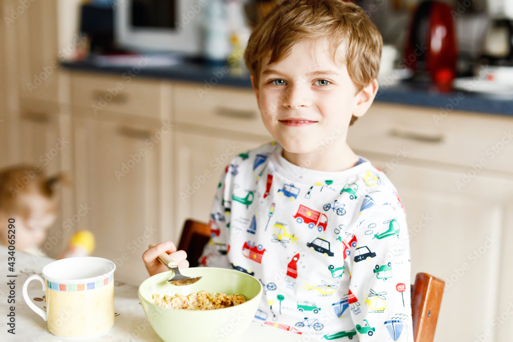 Happy little blond kid boy eating cereals for breakfast or lunch. Healthy eating for children in the morning. Child in colorful pajama nightwear having breakfast with milk and oat granola muesli Happy little blond kid boy eating cereals for breakfast or lunch. Healthy eating for children in the morning. Child in colorful pajama nightwear having breakfast with milk and oat granola muesli