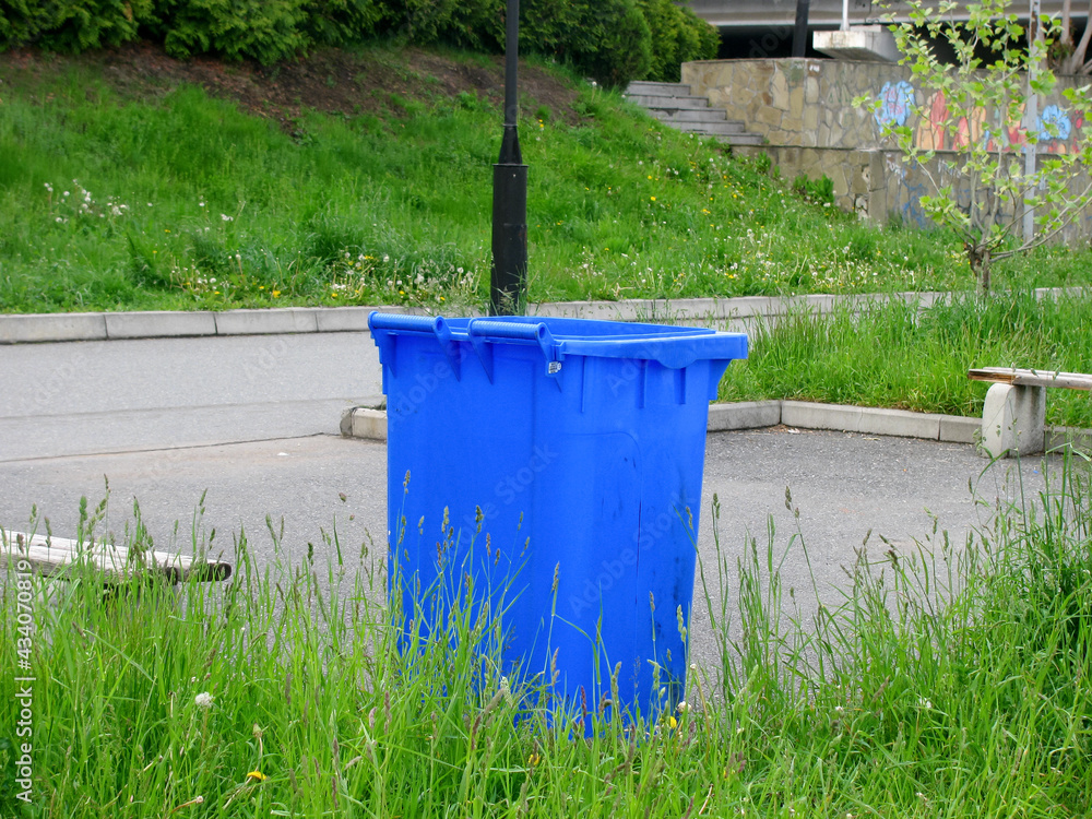 Blue dumpster on wheels on the street among the greenery in the park area. Blue dumpster on wheels on the street among the greenery in the park area.