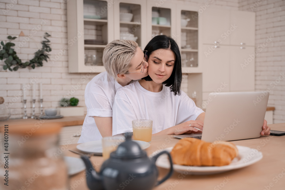 young woman kissing girlfriend while using laptop in kitchen young woman kissing girlfriend while using laptop in kitchen