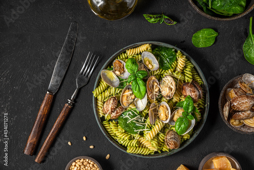 Seafood pasta with vongole clams, spinach, parmesan cheese, pine nuts and basil in a plate with fork and knife on black background