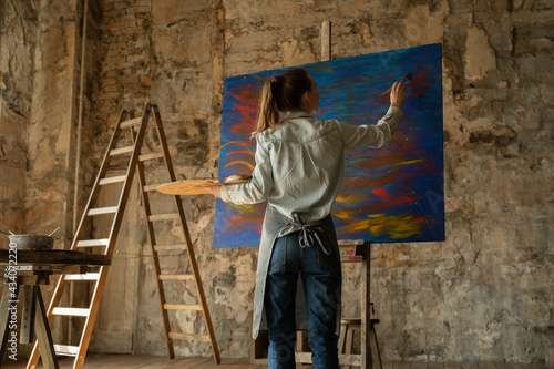 Female artist paints a picture of her studio. In her hands are brushes and a palette.  © Grustock