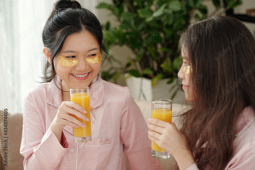 Attractive young smiling woman with undereye patches drinking glass of juice when spending time at home with her best friend Attractive young smiling woman with undereye patches drinking glass of juice when spending time at home with her best friend