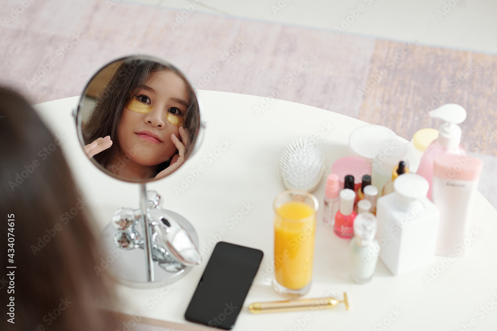 Pretty smiling young woman looking at magnifying mirror when applying undereye patches and lotions Pretty smiling young woman looking at magnifying mirror when applying undereye patches and lotions