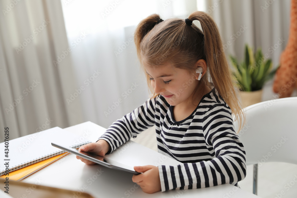 Adorable little girl doing homework with tablet and earphones at table indoors Adorable little girl doing homework with tablet and earphones at table indoors