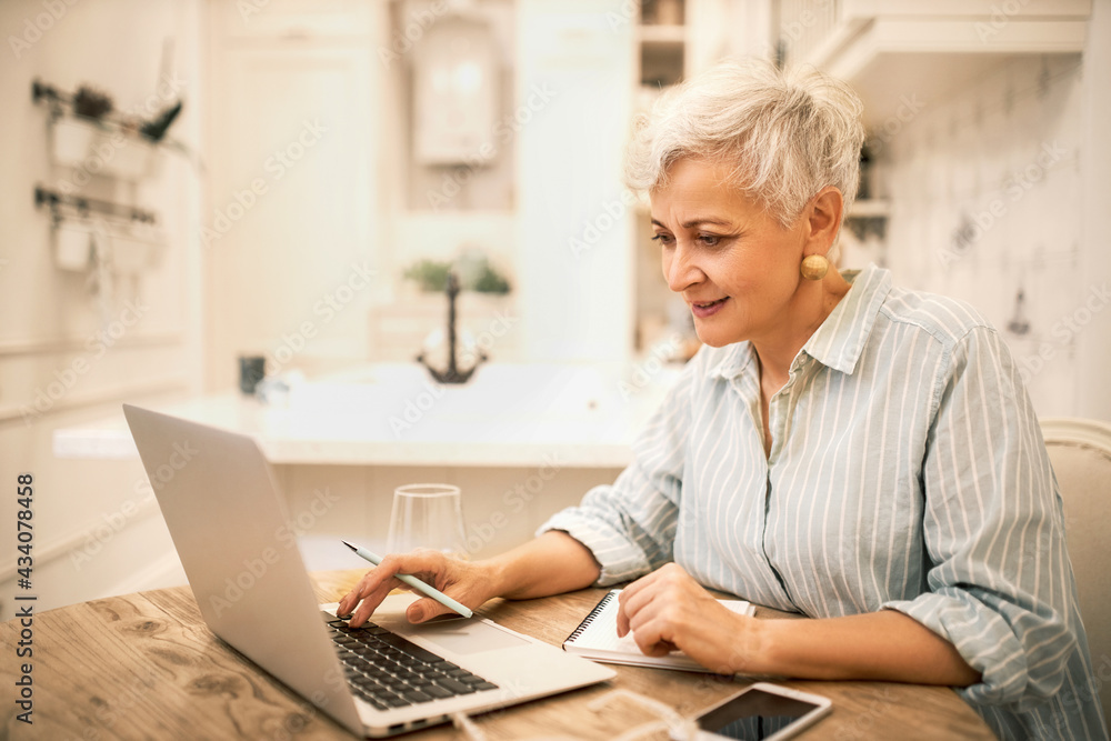 Portrait of focused stylish middle aged female using portable computer for distant work or online course, learning new marketing skills, making notes in copybook, having concentrated facial expression - obrazy, fototapety, plakaty 