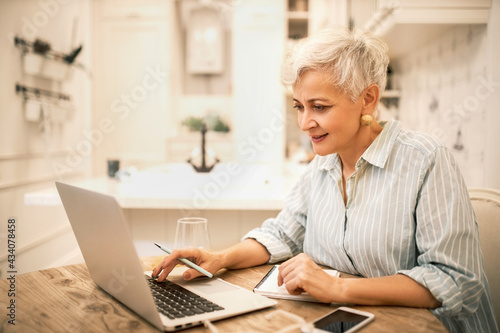 Portrait of focused stylish middle aged female using portable computer for distant work or online course, learning new marketing skills, making notes in copybook, having concentrated facial expression