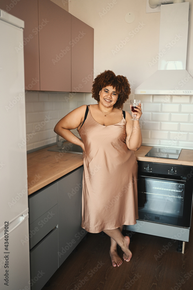 Beautiful curvy plus size African black woman afro hair drinking red wine on modern scandinavian style kitchen interior design. Body imperfection, body acceptance, body positive and diversity concept. Beautiful curvy plus size African black woman afro hair drinking red wine on modern scandinavian style kitchen interior design. Body imperfection, body acceptance, body positive and diversity concept.