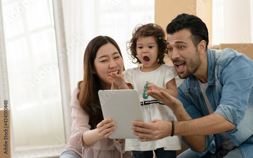 Happy young parents Caucasian father and Asian mother holding tablet with little kid girl show stone bracelet. They looking to device and showing excited expression face.