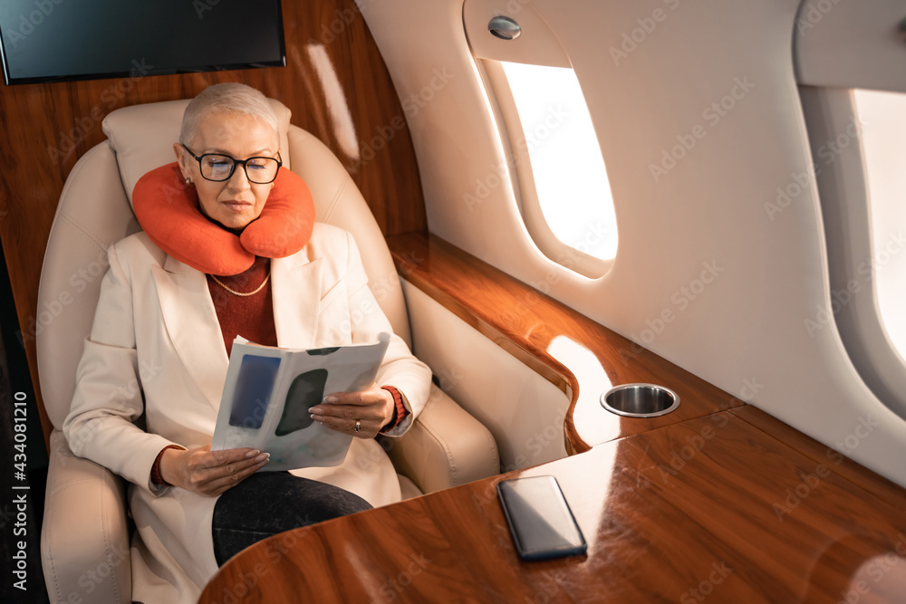 Businesswoman in neck pillow reading magazine near blurred smartphone in plane Businesswoman in neck pillow reading magazine near blurred smartphone in plane