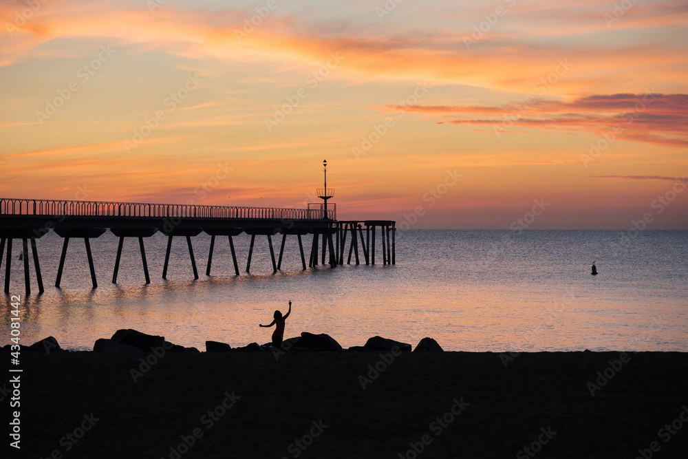 Silhouette of a woman dancing at the beach in the morning. Sunrise in the mediterranean sea with view to the pontoon. Pont del Petroli, Badalona, Barcelona, Catalonia, Spain.
