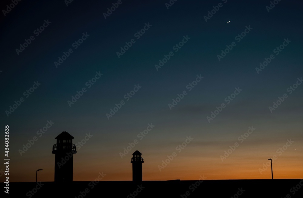 silhouette of lighthouse towers at a harbor with crescent moon in the evening