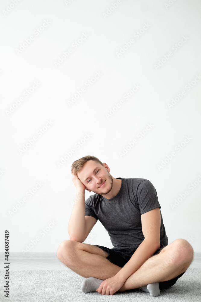 Positive man. Home relax. Lockdown lifestyle. Harmony peace. Handsome happy guy in t-shirt shorts sitting floor carpet in lotus pose leaning hand on leg light wall interior background copy space. Positive man. Home relax. Lockdown lifestyle. Harmony peace. Handsome happy guy in t-shirt shorts sitting floor carpet in lotus pose leaning hand on leg light wall interior background copy space.