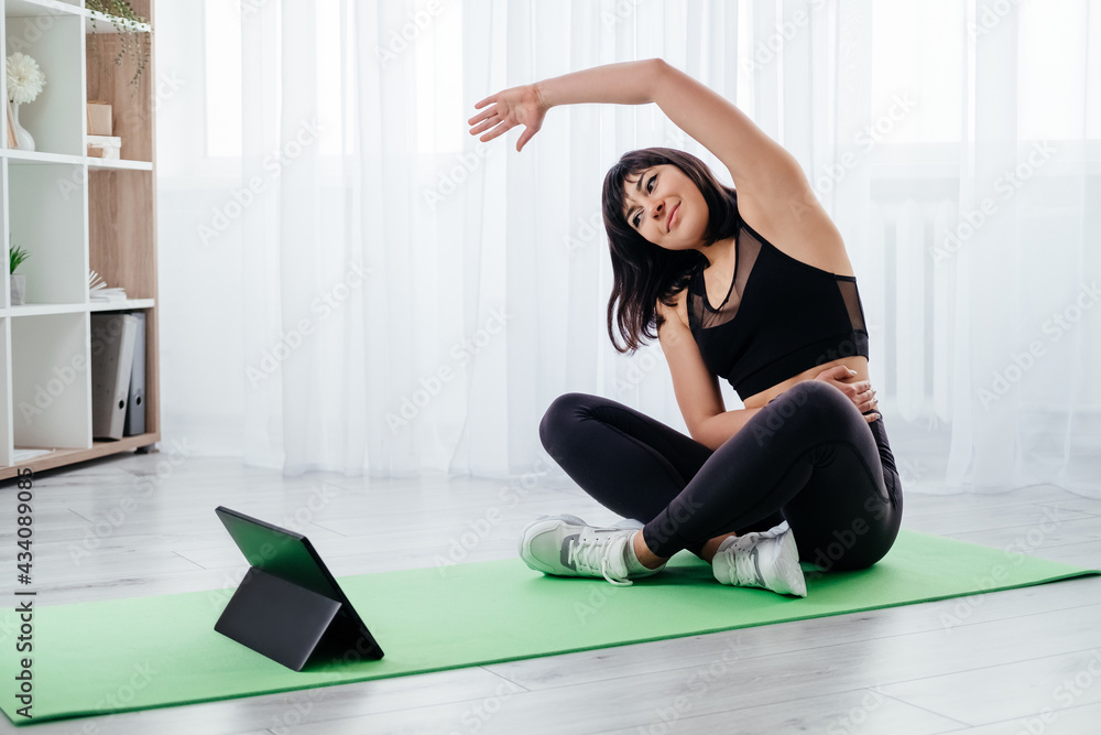 Fototapeta premium Online sport. Female fitness. Home training. Healthy body. Happy athletic woman in black sportswear doing stretching workout for hands waist before tablet computer light interior.