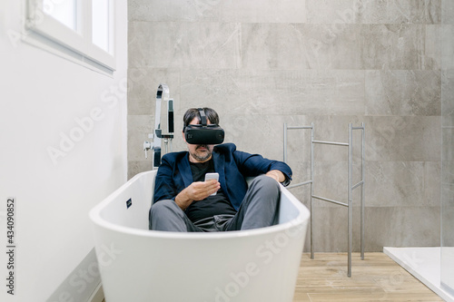 Fully clothed elegant adult male experiencing virtual reality in a bathtub as if he were on a tropical beach while messaging the smartphone. Virtual reality glasses. Virtual reality holiday.