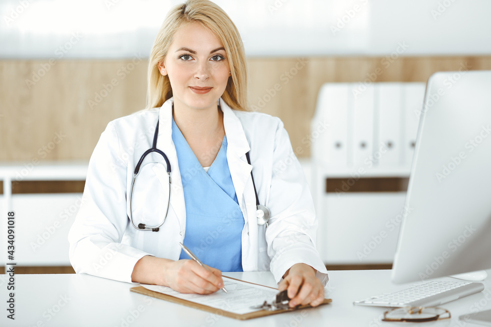 Portrate of woman-doctor at work while sitting at the desk in clinic. Blonde cheerful physician filling up medical form or prescription Portrate of woman-doctor at work while sitting at the desk in clinic. Blonde cheerful physician filling up medical form or prescription