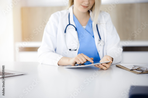 Unknown woman-doctor sitting in sunny clinic and using tablet pc, close-up. Data and best service in medicine