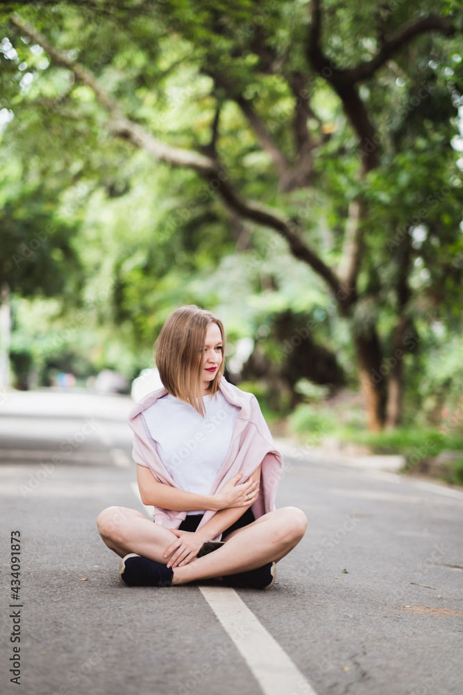 Young happy woman in white t-short is sitting in the middle of the asphalt road with greens on background Young happy woman in white t-short is sitting in the middle of the asphalt road with greens on background