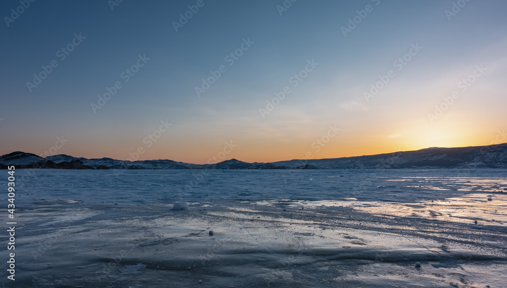 Sunset over the frozen lake plain. The sun is hidden behind a ridge, the sky is highlighted in orange. Reflections on ice. Baikal