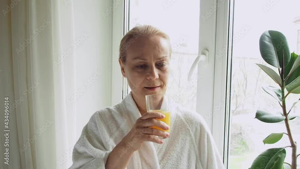 A mature woman in a white bathrobe drinks fruit juice while standing at the window at home. A mature woman in a white bathrobe drinks fruit juice while standing at the window at home.