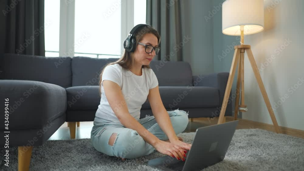 Casually dressed woman with headphones is sitting on carpet with laptop and working in cozy room. Remote work outside the office. Casually dressed woman with headphones is sitting on carpet with laptop and working in cozy room. Remote work outside the office.