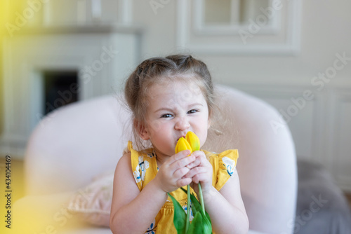 A two-year-old cute beautiful girl in a yellow suit sits on the sofa and holds yellow tulips in her hands, sniffs the flowers, wrinkles her nose.