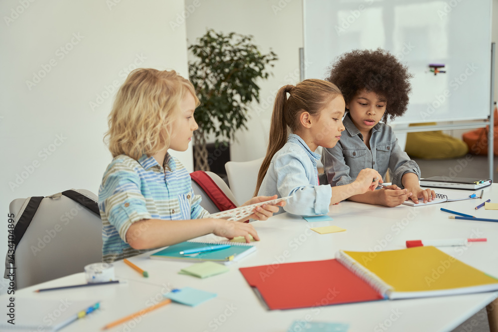 Cute diverse kids studying and drawing while sitting together at the table in elementary school classroom Cute diverse kids studying and drawing while sitting together at the table in elementary school classroom