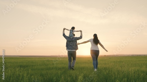Mom, dad and daughter are playing on the field, the child is sitting on dad's shoulders. Happy family runs in the park holding hands in the summer at sunset. Teamwork. Happy healthy childhood