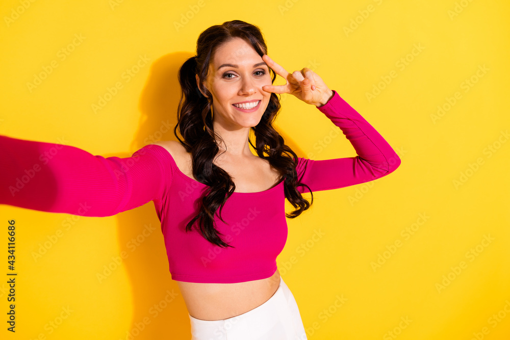 Self-portrait of attractive cheerful wavy-haired girl showing v-sign having fun isolated over bright yellow color background Self-portrait of attractive cheerful wavy-haired girl showing v-sign having fun isolated over bright yellow color background