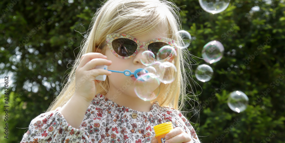 Portrait of fashionable toddler girl blowing soap bubbles in park.
Portrait of fashionable toddler girl blowing soap bubbles in park.