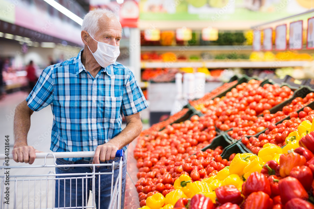 elderly caucasian man in mask with covid protection choosing tomatoes in vegetable section of supermarket