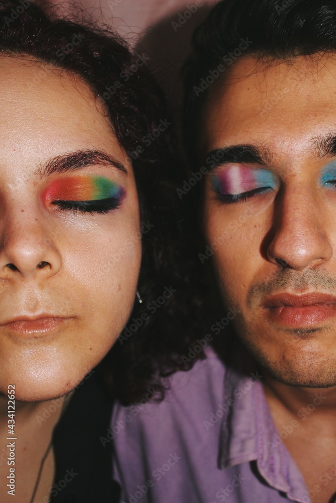 closeup portrait of queer young man & woman, friends of different ethnicity with lgbtqia+ nonbinary & transsexual pride flag eye makeup of spectrum colors - eyes closed