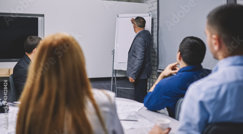 Experienced boss 50s coaching professional employees during involved briefing in board room  mature corporate director in formal suit teaching colleagues explaining business strategy near flipchart