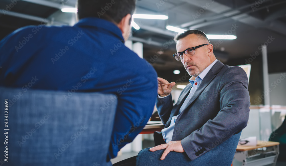 Caucaisan male boss in optical eyeglasses and formal suit explaining information to colleague discussing brainstorming process at desktop, businessmen in spectacles have conversation in office Caucaisan male boss in optical eyeglasses and formal suit explaining information to colleague discussing brainstorming process at desktop, businessmen in spectacles have conversation in office