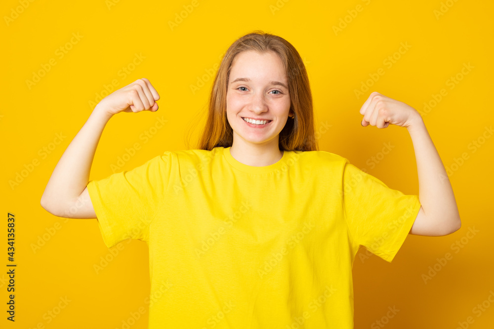Pretty teen girl keeps fit and healthy, raises hands and shows muscles, feels proud about her achievements in gym, smiles broadly, dressed in yellow casual wear, poses indoor shows real power. Pretty teen girl keeps fit and healthy, raises hands and shows muscles, feels proud about her achievements in gym, smiles broadly, dressed in yellow casual wear, poses indoor shows real power.