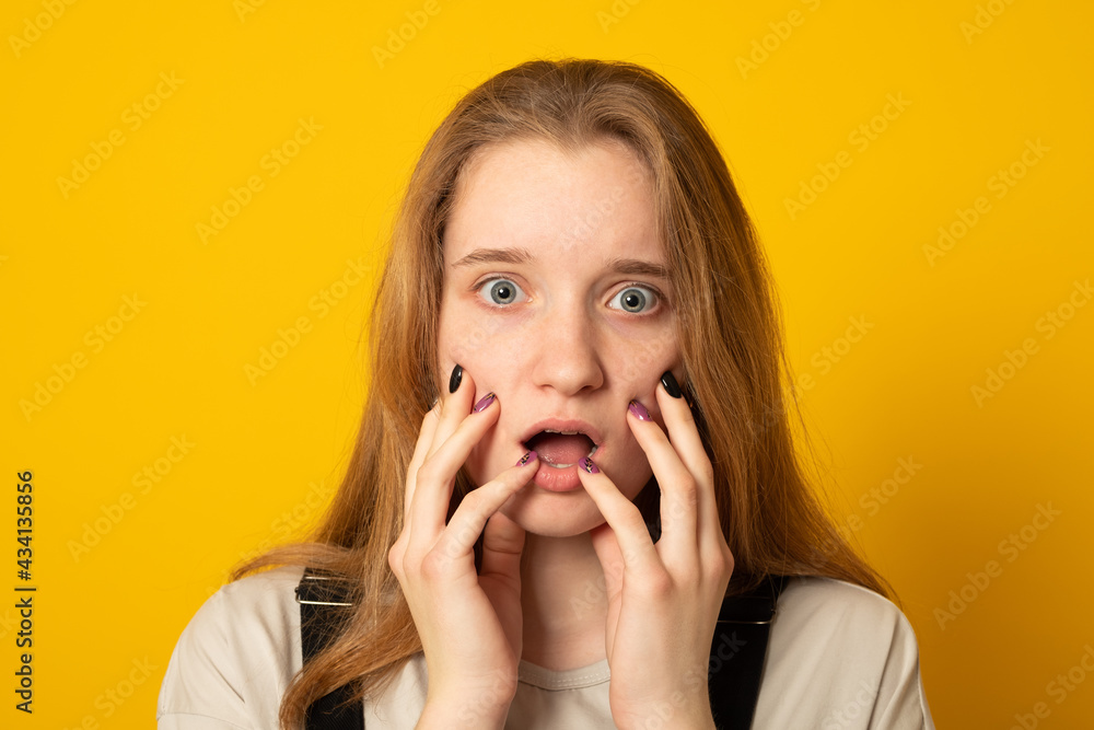 Terrified teen girl touches cheeks, keeps jaw dropped, frightened of spooky monster, isolated over yellow background. Concerned fearful girl grabs face. Terrified teen girl touches cheeks, keeps jaw dropped, frightened of spooky monster, isolated over yellow background. Concerned fearful girl grabs face.