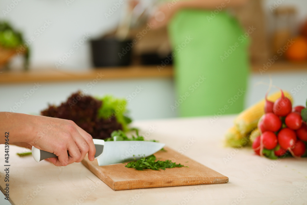 Unknown young woman slicing greens for a delicious fresh vegetarian salad while sitting and smiling at the kitchen desk, just hands, close-up. Cooking concept