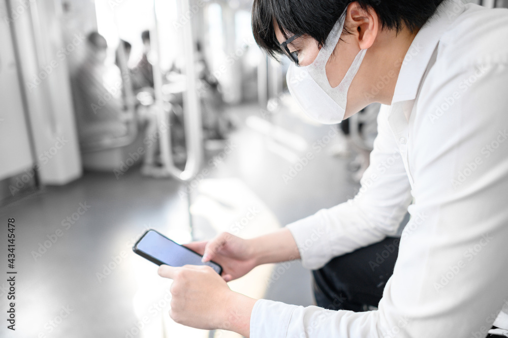Fototapeta premium Asian man wearing surgical face mask using smartphone on skytrain or urban train. Coronavirus (COVID-19) infection prevention in public transportation. Health awareness for pandemic protection