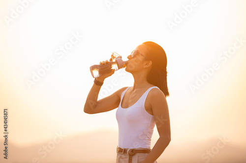 woman taking a break to drink from water bottle while hiking and poles standing on a rocky mountain ridge looking out valleys and peaks in a healthy outdoors lifestyle concept. explorer young woman.