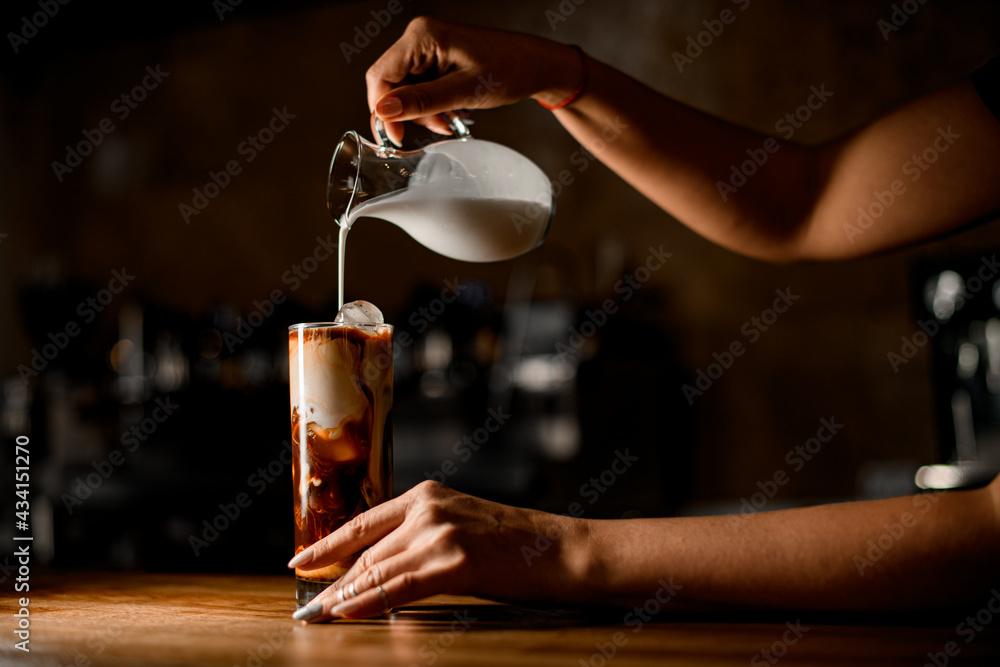 woman holds jug and carefully pours milk into glass with coffee and ice