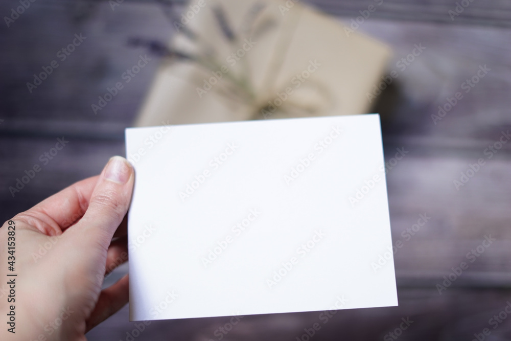 Fototapeta premium Woman's hand holding blank white card with a present on wooden background. Surprise gift mock-up scene. Empty space for text. Copy space.