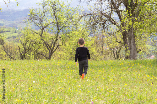 A small child with his back turned is playing in the meadow. He enjoys the beautiful nature and the view of the distant hills.