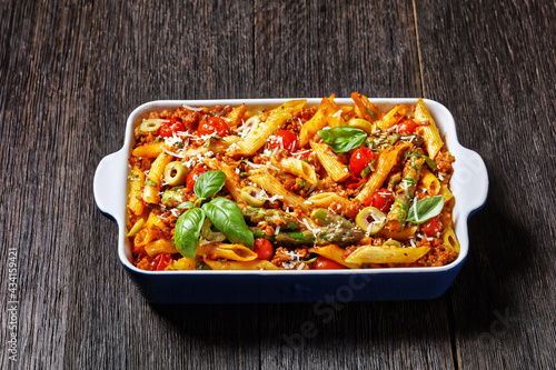 ground turkey penne pasta bake with asparagus, tomatoes and olives in a baking dish on a dark wooden table, italian cuisine