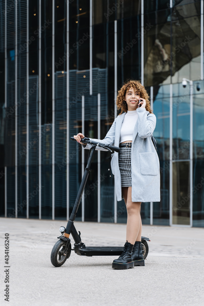 Full body of positive young African American female in blue coat having conversation via smartphone while standing on sidewalk with scooter Full body of positive young African American female in blue coat having conversation via smartphone while standing on sidewalk with scooter